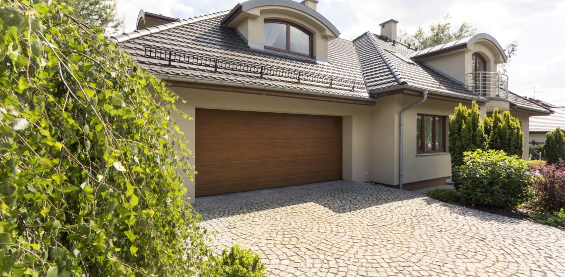 A contemporary beige house with a wooden garage door and tiled roof, surrounded by lush greenery and a cobblestone driveway.
