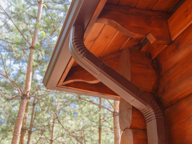 Close-up of a rustic wooden cabin's eaves with a gutter, set against a backdrop of pine trees.